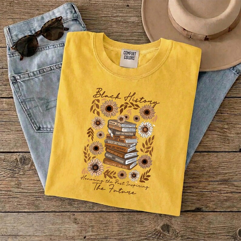 May include: Yellow t-shirt with the words "Black History" and a graphic of books and sunflowers. The shirt also has the text "Honoring the Past Inspiring The Future". The shirt is folded on top of blue jeans, sunglasses, and a hat.