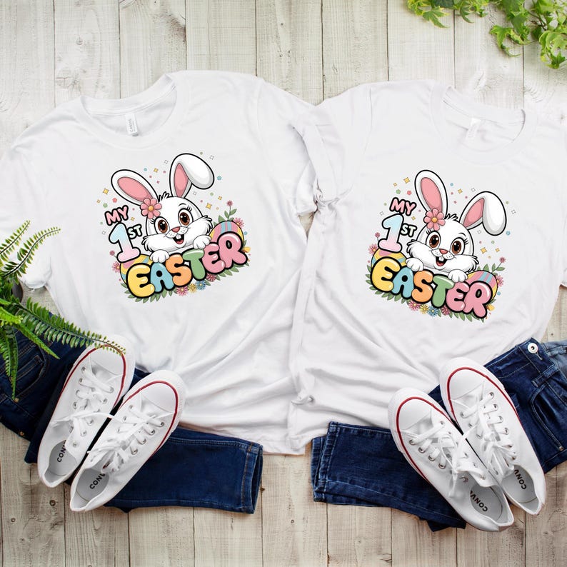 May include: Two white t-shirts with a cute cartoon bunny holding an Easter egg. The text on the shirts says "My 1st Easter".
