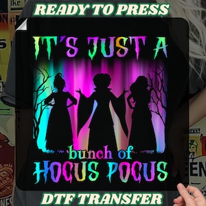May include: Black DTF transfer with the text "IT'S JUST A bunch of HOCUS POCUS". The design features silhouettes of three witches in front of a colorful background. The words "READY TO PRESS" and "DTF TRANSFER" are also included.