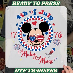 May include: A DTF transfer featuring Mickey Mouse wearing patriotic sunglasses, surrounded by red, white, and blue accents. The design includes the text "4th of July," "1776," and "Mickey Mouse," with fireworks and a rocket.