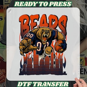 Peut inclure: Transfert DTF blanc avec un ours féroce en uniforme de football, tenant un ballon. L'ours est placé devant un paysage urbain et des flammes, avec le mot "BEARS" en orange. Les textes "READY TO PRESS" et "DTF TRANSFER" sont également présents.