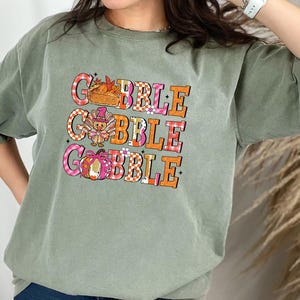 May include: Sage green t-shirt with the word "GOBBLE" in three lines. Each word is in a different color and pattern, with Thanksgiving-themed graphics. The shirt has short sleeves and a relaxed fit.