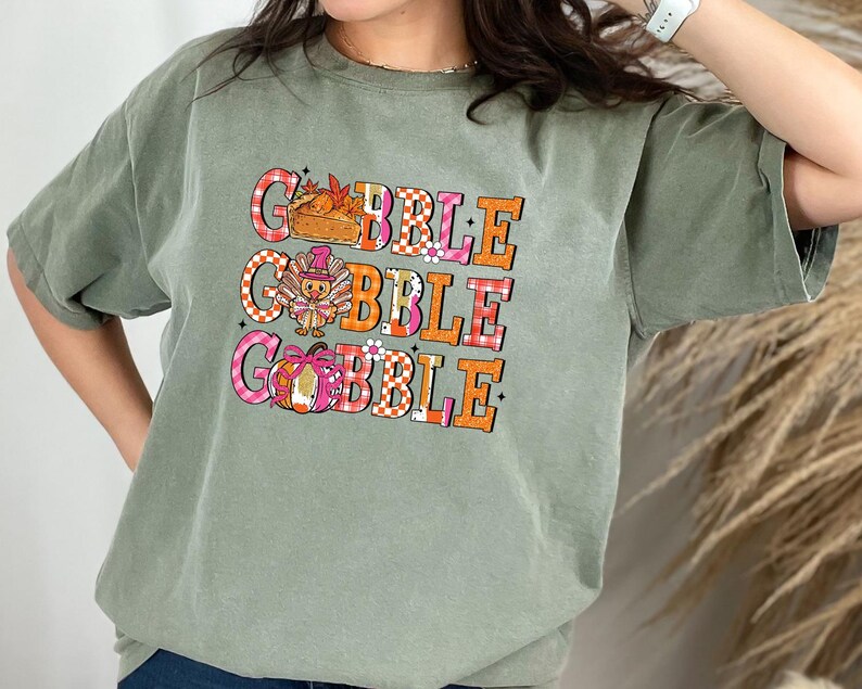 May include: Sage green t-shirt with the word "GOBBLE" in three lines. Each word is in a different color and pattern, with Thanksgiving-themed graphics. The shirt has short sleeves and a relaxed fit.