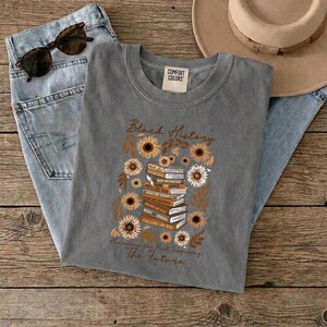 May include: A gray Comfort Colors t-shirt with the words "Black History" and a design of sunflowers and books. The text "Honoring the Past, Inspiring The Future" is also printed on the shirt. Sunglasses and jeans are in the background.
