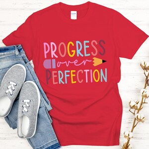 May include: A red t-shirt with the words "PROGRESS over PERFECTION" in colorful letters and a pencil graphic. The shirt is paired with blue jeans and gray sneakers with white laces. A sprig of cotton stems is on the right.
