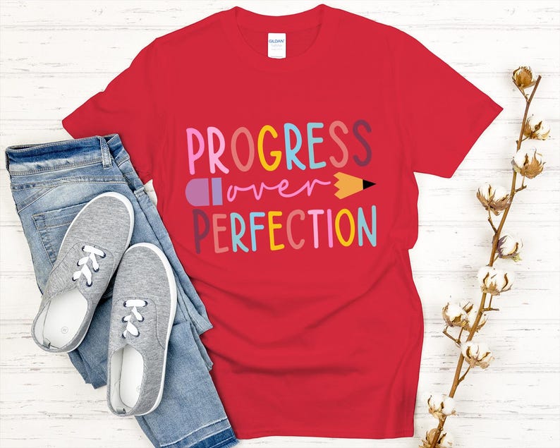 May include: A red t-shirt with the words "PROGRESS over PERFECTION" in colorful letters and a pencil graphic. The shirt is paired with blue jeans and gray sneakers with white laces. A sprig of cotton stems is on the right.