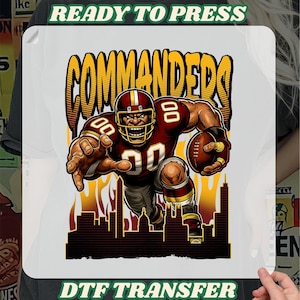 May include: A DTF transfer featuring a muscular football player in a burgundy and gold uniform, holding a football. The word "COMMANDERS" is in large, yellow, outlined letters. The image includes a cityscape silhouette and the text "READY TO PRESS" and "DTF TRANSFER".