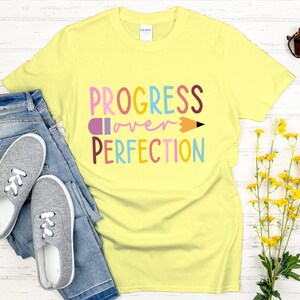 May include: A light yellow t-shirt with the words "PROGRESS over PERFECTION" in colorful letters and a pencil graphic. The shirt is paired with blue jeans and gray sneakers. Sunglasses and yellow flowers are also in the image.