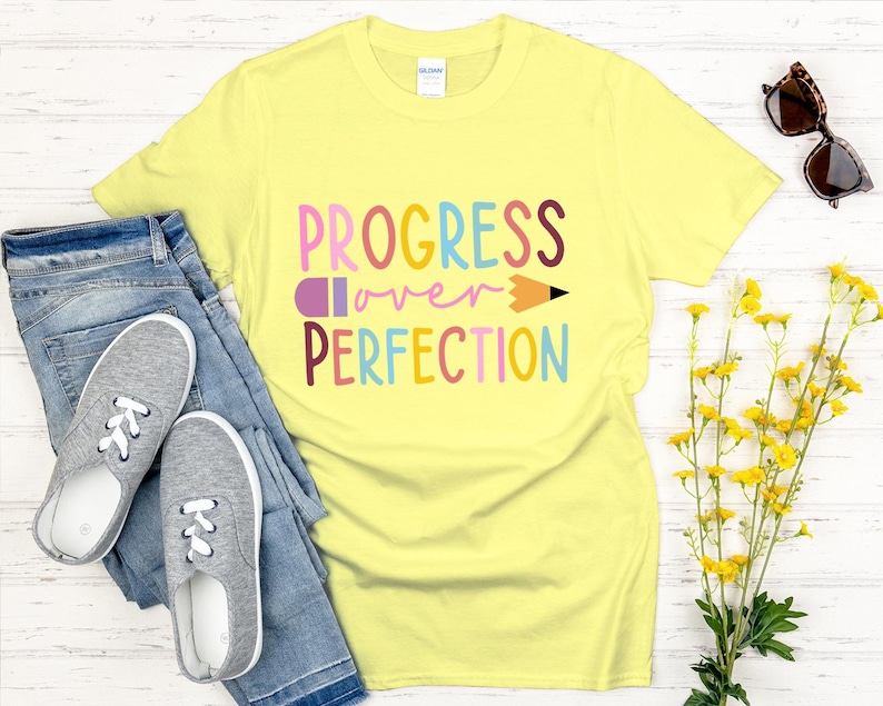 May include: A light yellow t-shirt with the words "PROGRESS over PERFECTION" in colorful letters and a pencil graphic. The shirt is paired with blue jeans and gray sneakers. Sunglasses and yellow flowers are also in the image.