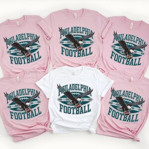 Może przedstawiać: Kolekcja r&oacute;żowych i białych koszulek z motywem futbolu amerykańskiego. Koszulki mają napis "PHILADELPHIA FOOTBALL" z grafiką orła. Projekt zawiera piłkę nożną i rok "1933".