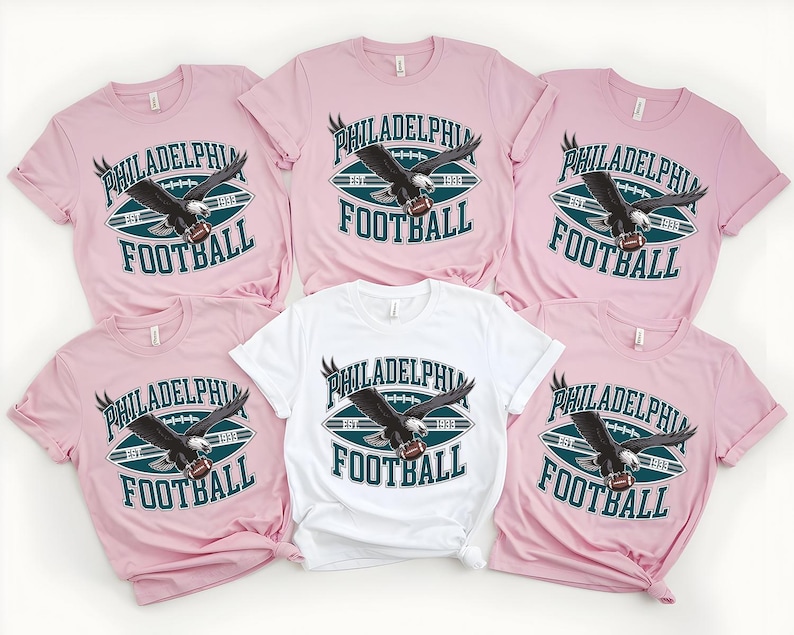 Może przedstawiać: Kolekcja r&oacute;żowych i białych koszulek z motywem futbolu amerykańskiego. Koszulki mają napis "PHILADELPHIA FOOTBALL" z grafiką orła. Projekt zawiera piłkę nożną i rok "1933".