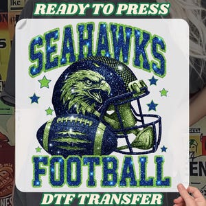 May include: A white DTF transfer featuring a football helmet, football, and eagle graphic. The words "SEAHAWKS FOOTBALL" are in blue glitter text with green accents. The text "READY TO PRESS" and "DTF TRANSFER" are also included.