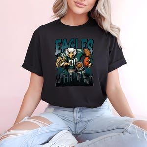 May include: Black t-shirt with a cartoon eagle in a football uniform, holding a football. The word "EAGLES" is printed above the eagle in teal and white. A cityscape silhouette is at the bottom of the design.
