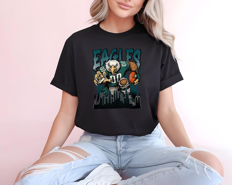 May include: Black t-shirt with a cartoon eagle in a football uniform, holding a football. The word "EAGLES" is printed above the eagle in teal and white. A cityscape silhouette is at the bottom of the design.