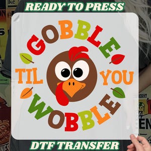 May include: A DTF transfer with a Thanksgiving theme. The design features a cartoon turkey with the words "GOBBLE TIL YOU WOBBLE" in various colors. The text "READY TO PRESS" and "DTF TRANSFER" are also included.