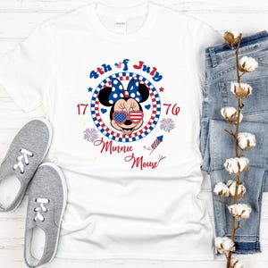 May include: White t-shirt featuring a Minnie Mouse design with patriotic elements. The design includes Minnie wearing sunglasses with a stars and stripes pattern, a blue bow, and the text "4th of July 1776 Minnie Mouse". Gray sneakers and blue jeans are also in the image.