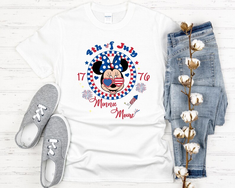 May include: White t-shirt featuring a Minnie Mouse design with patriotic elements. The design includes Minnie wearing sunglasses with a stars and stripes pattern, a blue bow, and the text "4th of July 1776 Minnie Mouse". Gray sneakers and blue jeans are also in the image.