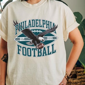 Może przedstawiać: Kremowa koszulka z grafiką orła trzymającego piłkę do futbolu amerykańskiego. Projekt zawiera napisy "PHILADELPHIA FOOTBALL" i "EST. 1933" w kolorze turkusowym i białym, w stylu vintage sportowym.