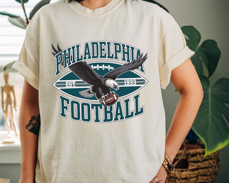 Może przedstawiać: Kremowa koszulka z grafiką orła trzymającego piłkę do futbolu amerykańskiego. Projekt zawiera napisy "PHILADELPHIA FOOTBALL" i "EST. 1933" w kolorze turkusowym i białym, w stylu vintage sportowym.