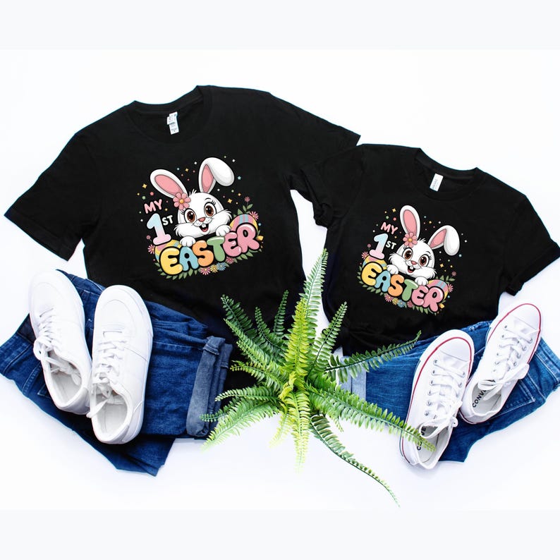May include: Two black t-shirts with a white bunny rabbit graphic and the text "My 1st Easter". The shirts are laid out on a white surface with blue jeans and white sneakers.
