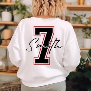 May include: White sweatshirt with a large black number 7 outlined in red on the back. Below the number is the name "Smith" in a cursive font. The sweatshirt is a crew neck style.