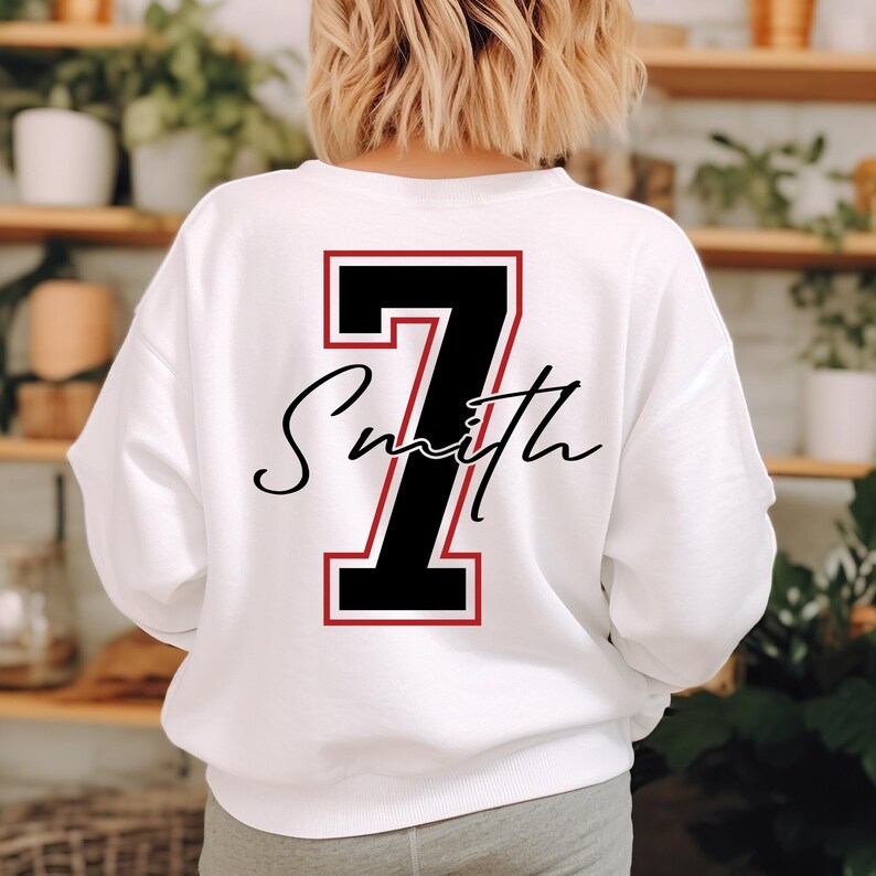 May include: White sweatshirt with a large black number 7 outlined in red on the back. Below the number is the name "Smith" in a cursive font. The sweatshirt is a crew neck style.