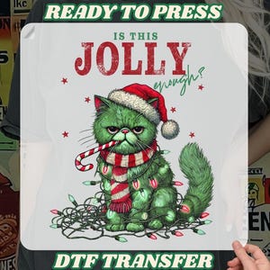 May include: A DTF transfer featuring a grumpy green cat wearing a Santa hat, a red and white scarf, and wrapped in Christmas lights. The text "IS THIS JOLLY enough?" is above the cat, with "READY TO PRESS" and "DTF TRANSFER" at the top and bottom.