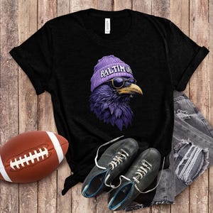 Pu&ograve; includere: T-shirt nero con un corvo viola che indossa un berretto viola con la scritta "Baltimore" e occhiali da sole. Un pallone da football americano marrone e scarpe nere sono accanto alla maglietta. La maglietta &egrave; su una superficie di legno.