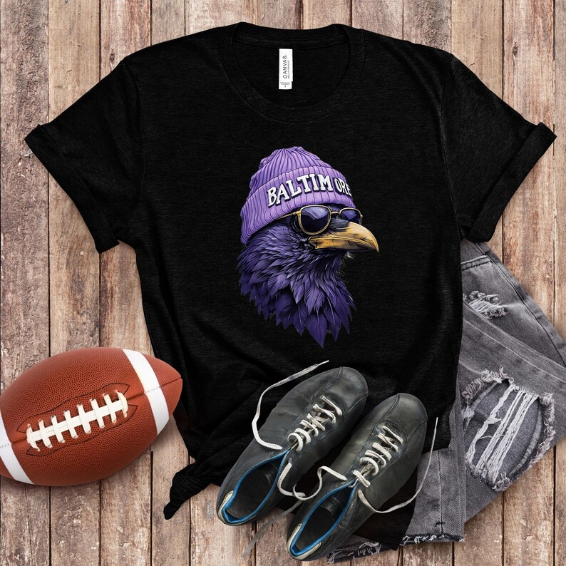 Pu&ograve; includere: T-shirt nero con un corvo viola che indossa un berretto viola con la scritta "Baltimore" e occhiali da sole. Un pallone da football americano marrone e scarpe nere sono accanto alla maglietta. La maglietta &egrave; su una superficie di legno.