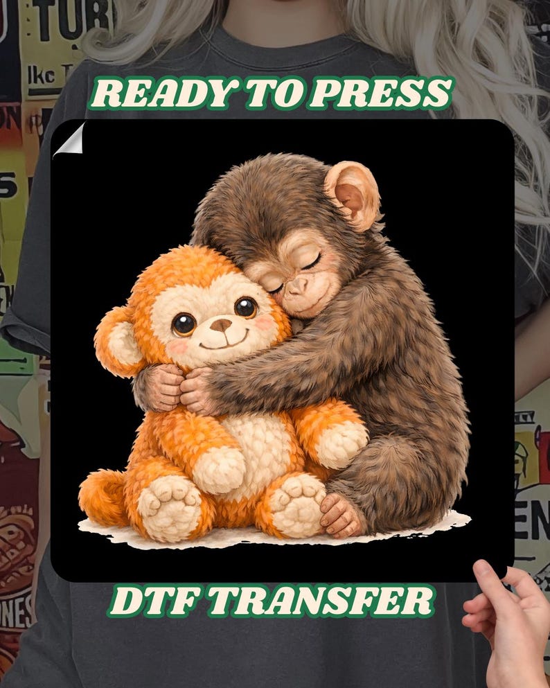 Op de afbeelding: Een DTF-transfer met een bruine aap die een oranje pluche aap omhelst. De bruine aap heeft zijn ogen gesloten en de oranje aap heeft grote, vriendelijke ogen. De woorden "READY TO PRESS" en "DTF TRANSFER" staan op de afbeelding.