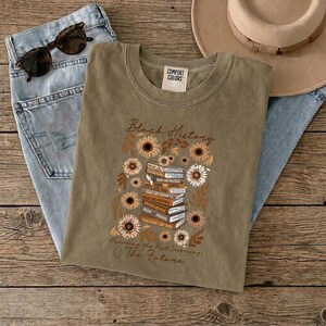 May include: An olive green t-shirt with the words "Black History" and "Honoring the Past, Inspiring the Future" printed on it. The design includes a stack of books surrounded by sunflowers and other floral elements. Sunglasses and jeans are also in the image.