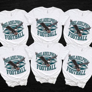 Może przedstawiać: Sześć białych koszulek z motywem futbolu amerykańskiego Philadelphia Eagles. Projekt przedstawia ciemnego orła trzymającego piłkę do futbolu, ze słowami "PHILADELPHIA FOOTBALL" w kolorze morskim i białym.