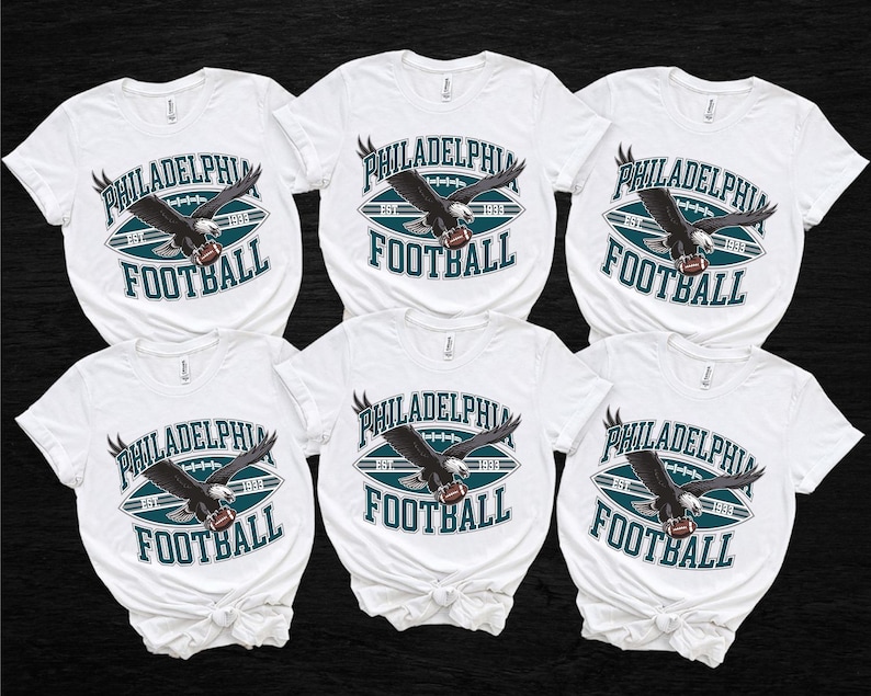 Może przedstawiać: Sześć białych koszulek z motywem futbolu amerykańskiego Philadelphia Eagles. Projekt przedstawia ciemnego orła trzymającego piłkę do futbolu, ze słowami "PHILADELPHIA FOOTBALL" w kolorze morskim i białym.