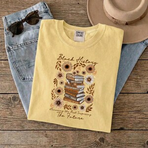May include: A yellow t-shirt with the words "Black History" and a stack of books surrounded by sunflowers. The text "Honoring the Past Inspiring The Future" is below the graphic. Sunglasses and jeans are in the background.