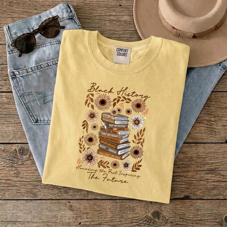 May include: A yellow t-shirt with the words "Black History" and a stack of books surrounded by sunflowers. The text "Honoring the Past Inspiring The Future" is below the graphic. Sunglasses and jeans are in the background.