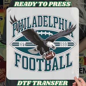 Może przedstawiać: Transfer DTF z napisem "PHILADELPHIA FOOTBALL" i grafiką orła trzymającego piłkę nożną. Projekt zawiera tekst "EST. 1933" oraz "READY TO PRESS" i "DTF TRANSFER" w kolorze zielonym.