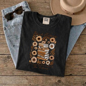 May include: Black t-shirt with a sunflower and book design. The shirt features the words "Black History" and "Inspiring The Future". Sunglasses and blue jeans are also in the image.