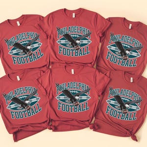 Może przedstawiać: Sześć czerwonych koszulek z napisem "PHILADELPHIA FOOTBALL" i grafiką orła. Tekst jest biały i morski, na tle kształtu piłki nożnej w kolorze morskim i białym. Orzeł jest czarno-biały.