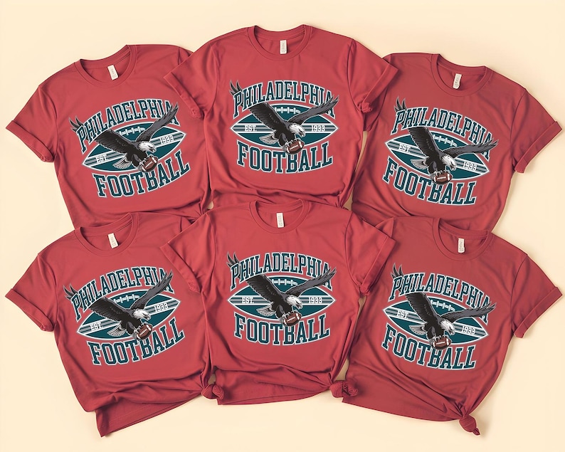 Może przedstawiać: Sześć czerwonych koszulek z napisem "PHILADELPHIA FOOTBALL" i grafiką orła. Tekst jest biały i morski, na tle kształtu piłki nożnej w kolorze morskim i białym. Orzeł jest czarno-biały.