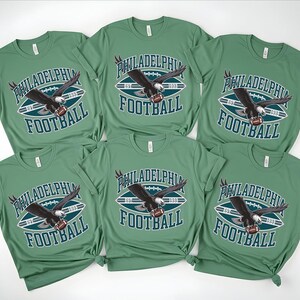 Może przedstawiać: Sześć zielonych koszulek z napisem "PHILADELPHIA FOOTBALL" i grafiką orła trzymającego piłkę do futbolu amerykańskiego. Koszulki są z kr&oacute;tkim rękawem i mają okrągły dekolt. Grafika jest w kolorze białym, morskim i czarnym.