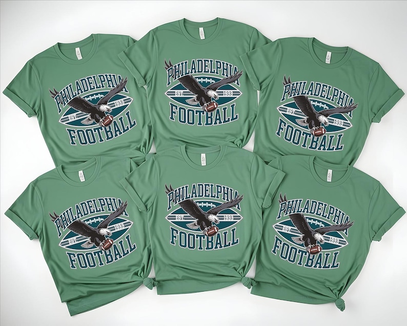 Może przedstawiać: Sześć zielonych koszulek z napisem "PHILADELPHIA FOOTBALL" i grafiką orła trzymającego piłkę do futbolu amerykańskiego. Koszulki są z kr&oacute;tkim rękawem i mają okrągły dekolt. Grafika jest w kolorze białym, morskim i czarnym.