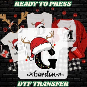 May include: A Christmas-themed DTF transfer design featuring a large black letter "G" adorned with a Santa hat and string lights. The design includes the name "Gordon" and is displayed on white t-shirts and a red jacket.