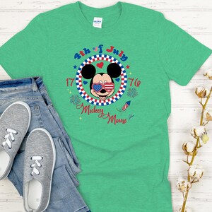 May include: A green t-shirt featuring a graphic of Mickey Mouse wearing American flag sunglasses, with the text "4th of July 1776 Mickey Mouse". The shirt is paired with blue jeans and gray slip-on shoes.