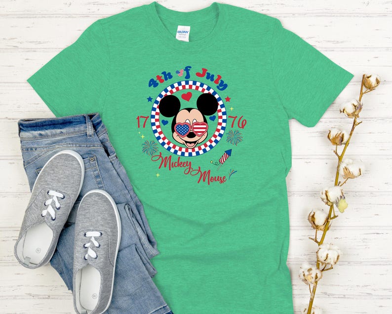 May include: A green t-shirt featuring a graphic of Mickey Mouse wearing American flag sunglasses, with the text "4th of July 1776 Mickey Mouse". The shirt is paired with blue jeans and gray slip-on shoes.
