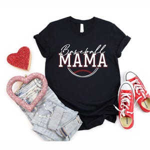 May include: Black t-shirt with the text "Baseball MAMA" in white and red lettering, with a baseball graphic. The image also includes a pair of blue jeans, red sneakers, and heart-shaped decorations.