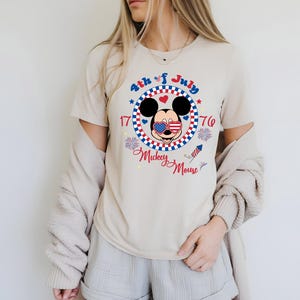 May include: Beige t-shirt featuring a patriotic Mickey Mouse design for the 4th of July. The graphic includes Mickey with American flag sunglasses, surrounded by a red, white, and blue circle with the text "4th of July 1776 Mickey Mouse."