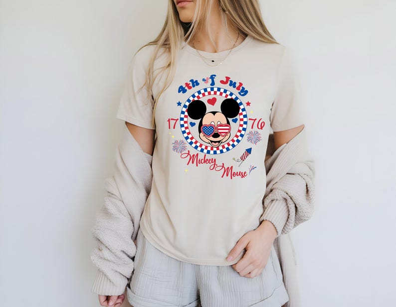 May include: Beige t-shirt featuring a patriotic Mickey Mouse design for the 4th of July. The graphic includes Mickey with American flag sunglasses, surrounded by a red, white, and blue circle with the text "4th of July 1776 Mickey Mouse."