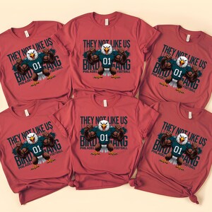 K&ouml;nnte beinhalten: Rote T-Shirts mit einer Grafik eines Adlers, der ein Football-Trikot mit der Nummer 01 tr&auml;gt. Auf den Shirts steht "THEY NOT LIKE US BIRD GANG PHILADELPHIA FOOTBALL."