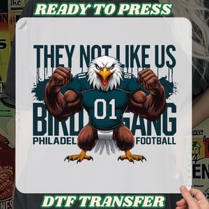 K&ouml;nnte beinhalten: DTF-Transfer mit einem muskul&ouml;sen Adler, der ein t&uuml;rkisfarbenes Football-Trikot mit der Nummer 01 tr&auml;gt. Der Text lautet "THEY NOT LIKE US" und "BIRD GANG PHILADELPHIA FOOTBALL". Die W&ouml;rter "READY TO PRESS" und "DTF TRANSFER" sind ebenfalls vorhanden.
