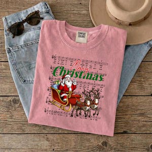 May include: A pink t-shirt featuring a graphic of Santa Claus in a red sleigh pulled by two reindeer over a background of musical sheet notes. The text Merry Christmas is printed in green script above the illustration. The shirt has a Comfort Colors label.
